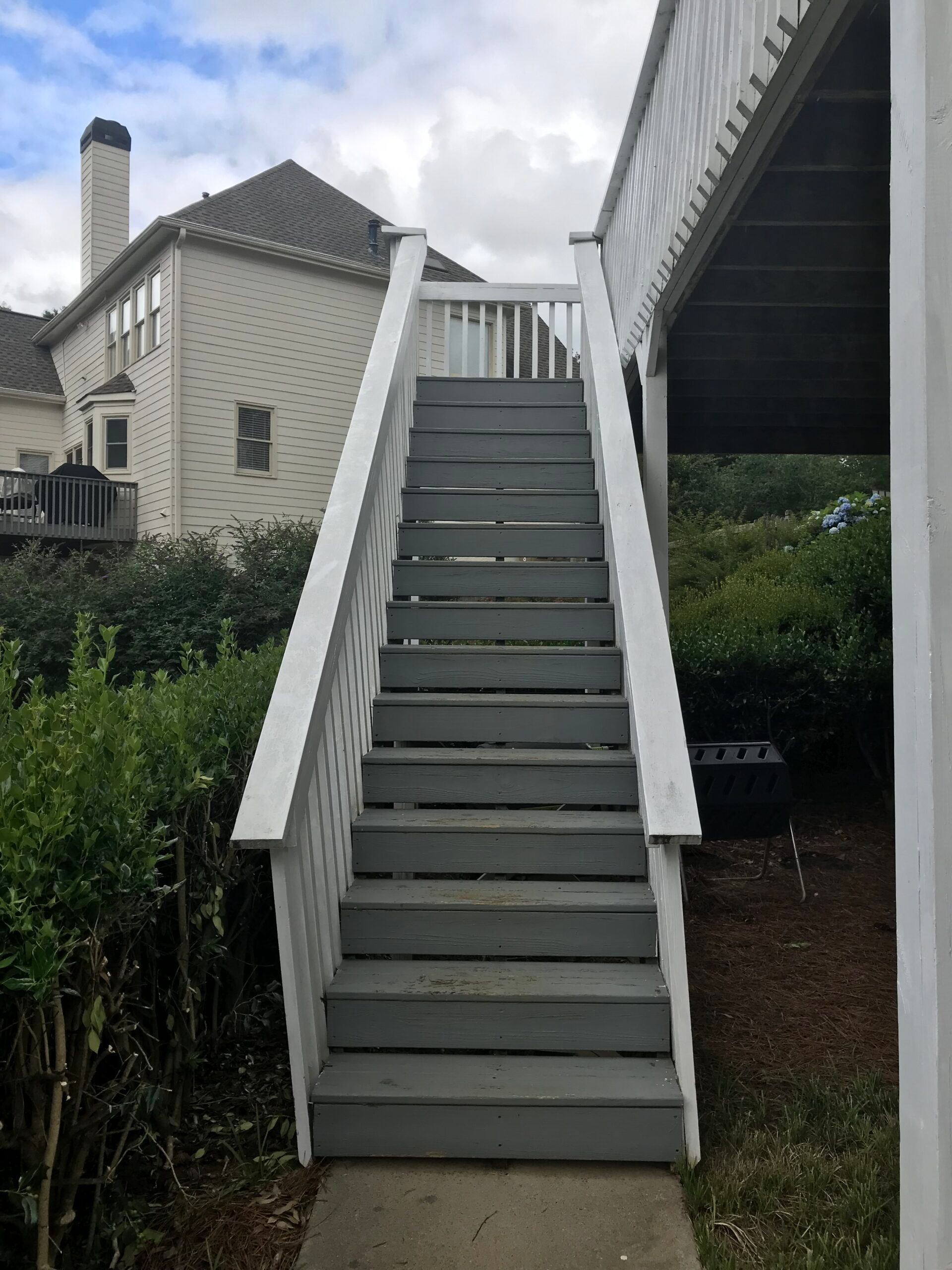 A set of grey outdoor wooden stairs with white railings leading up to a raised deck in a residential backyard.