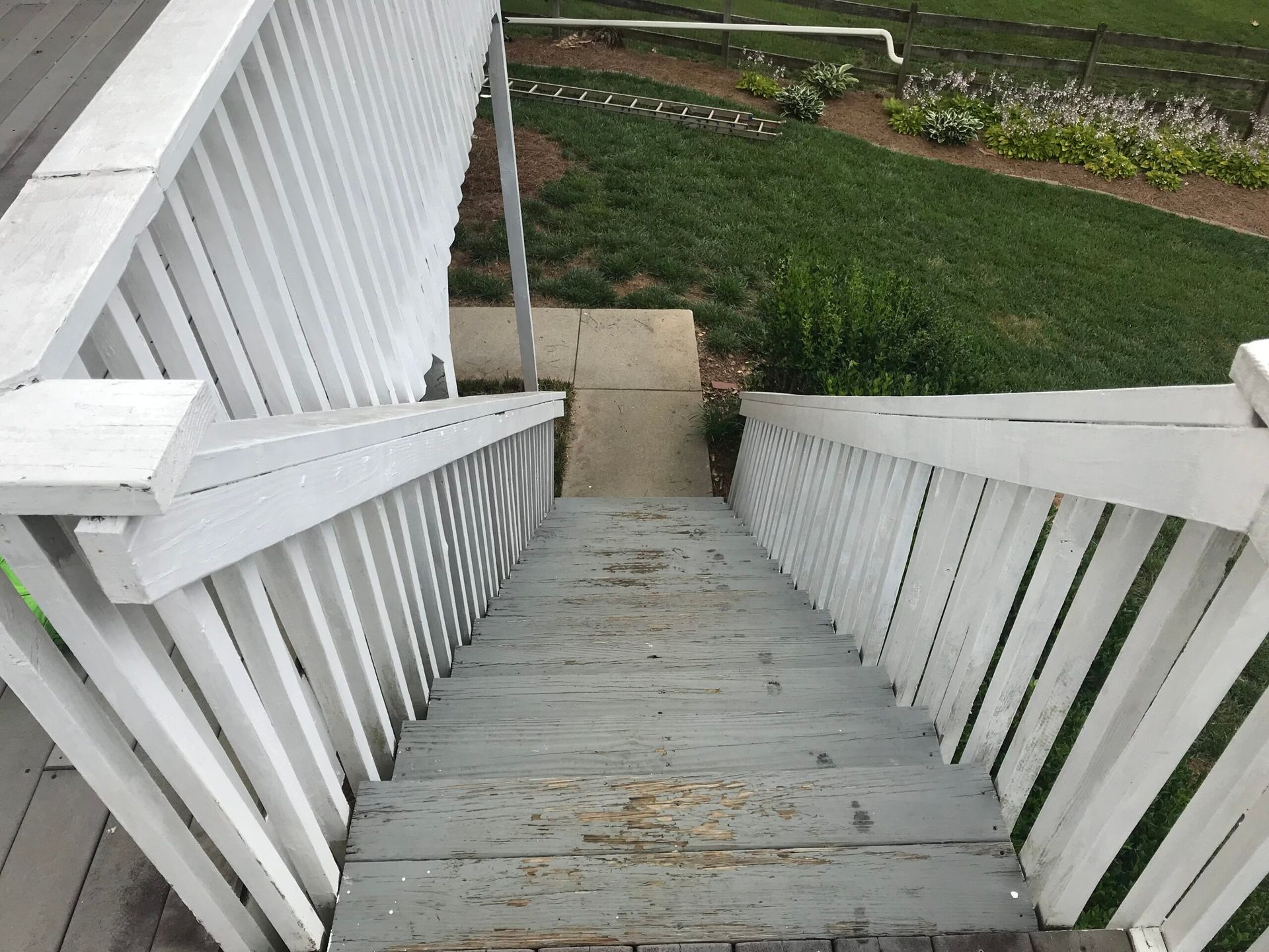 A high-angle view looking down a set of weathered grey wooden outdoor stairs with white railings, leading to a concrete path.