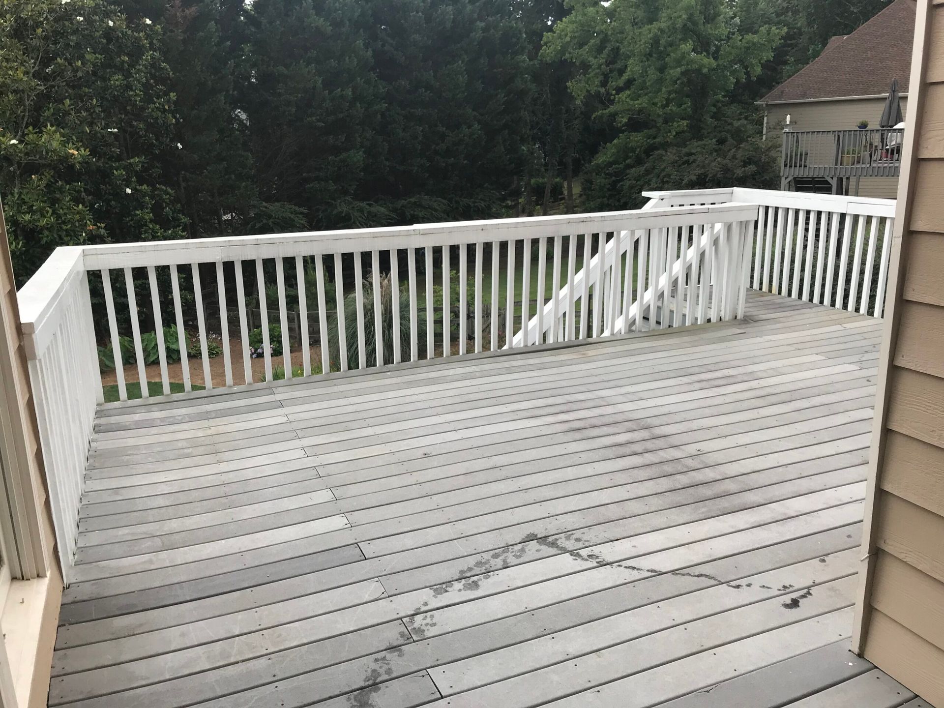 A weathered grey wooden deck with white railing, viewed from a doorway, looking out toward trees and a neighboring house.