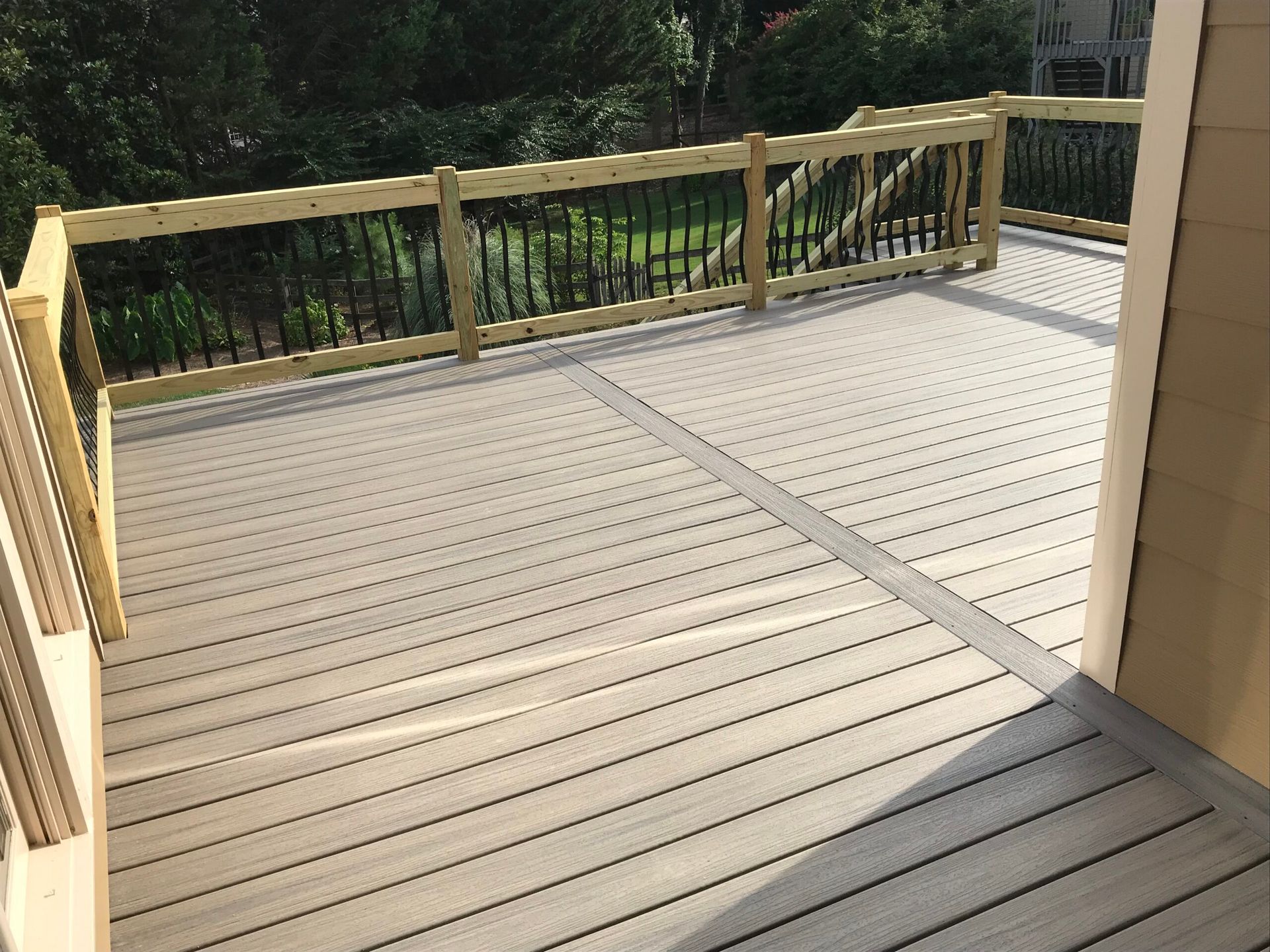 A gray composite deck with a light-colored central border and wooden railing overlooking a green, tree-lined yard.