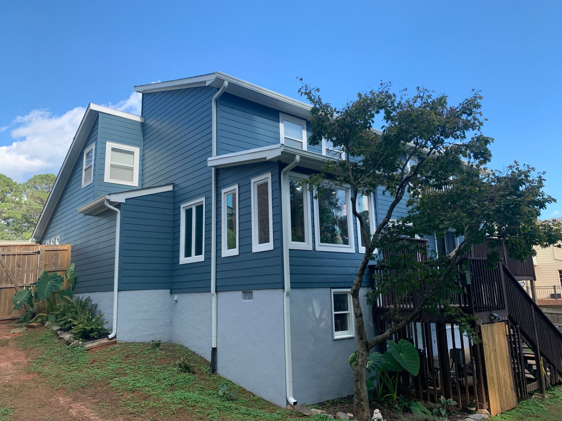 A multi-level house with dark blue horizontal siding and a light grey foundation, featuring a wooden deck and a tree.