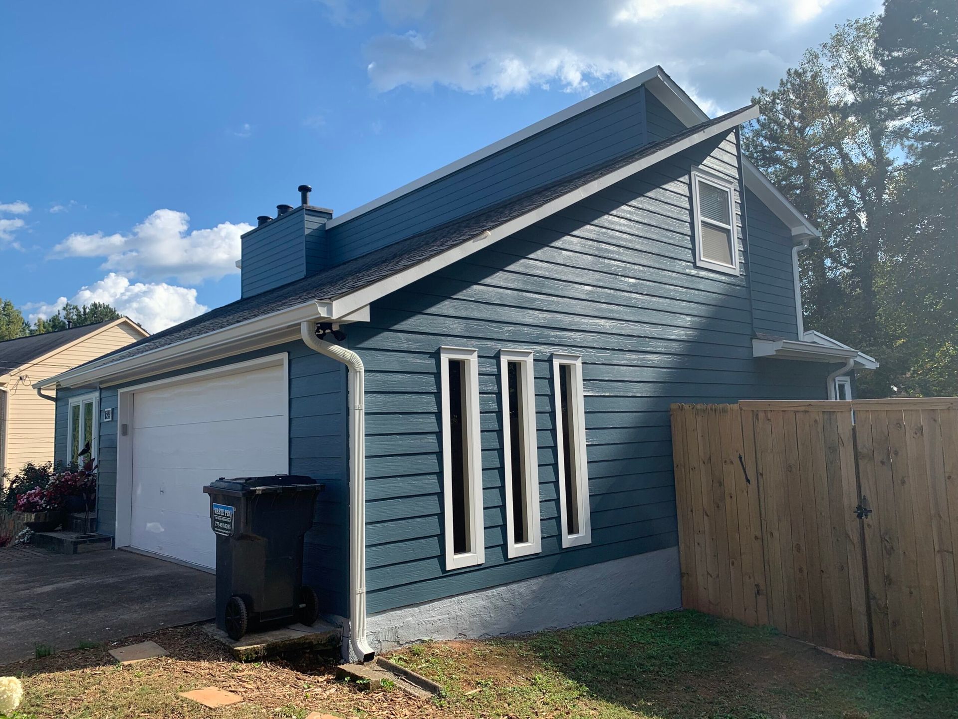 A side view of a blue, two-story house with a white garage door, three narrow windows, and a wooden fence.