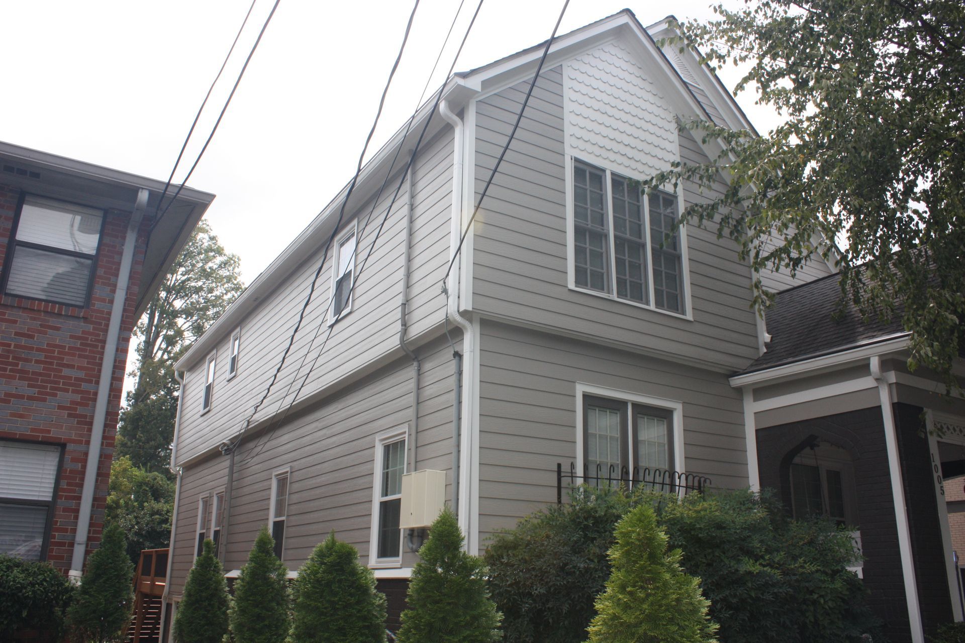 A side view of a light-gray, two-story residential house with white trim, set behind a row of small evergreen bushes.