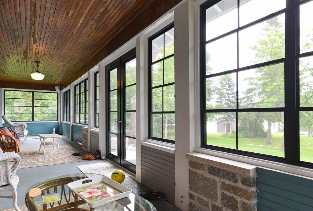 A sunroom with a wood-paneled ceiling, large black-framed windows, stone walls, and wicker furniture.
