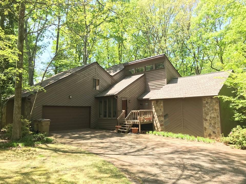 A brown, multi-level house with a wooden staircase entrance, set in a lush, wooded environment with a paved driveway.