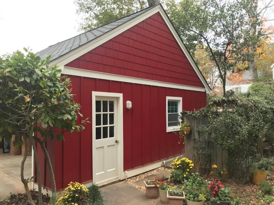 A red shed with white trim, a single door with window panes, and a small window, surrounded by greenery and potted plants.