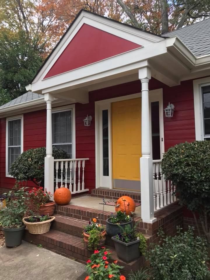 A red house with a bright yellow front door, white porch columns, brick steps, and potted plants with orange pumpkins.