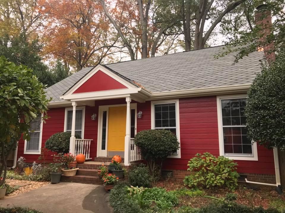 A single-story house with bright red siding, a yellow front door, and a white-pillared porch, surrounded by fall foliage.