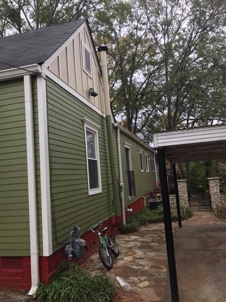Side view of a green-sided house with a white trim, a red brick base, a tall chimney, and a bike leaning against the wall.