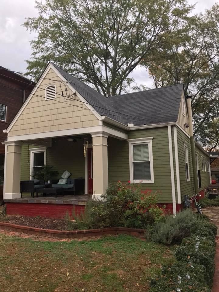 An olive green craftsman bungalow with a beige gabled porch and red foundation, surrounded by trees and landscaping.