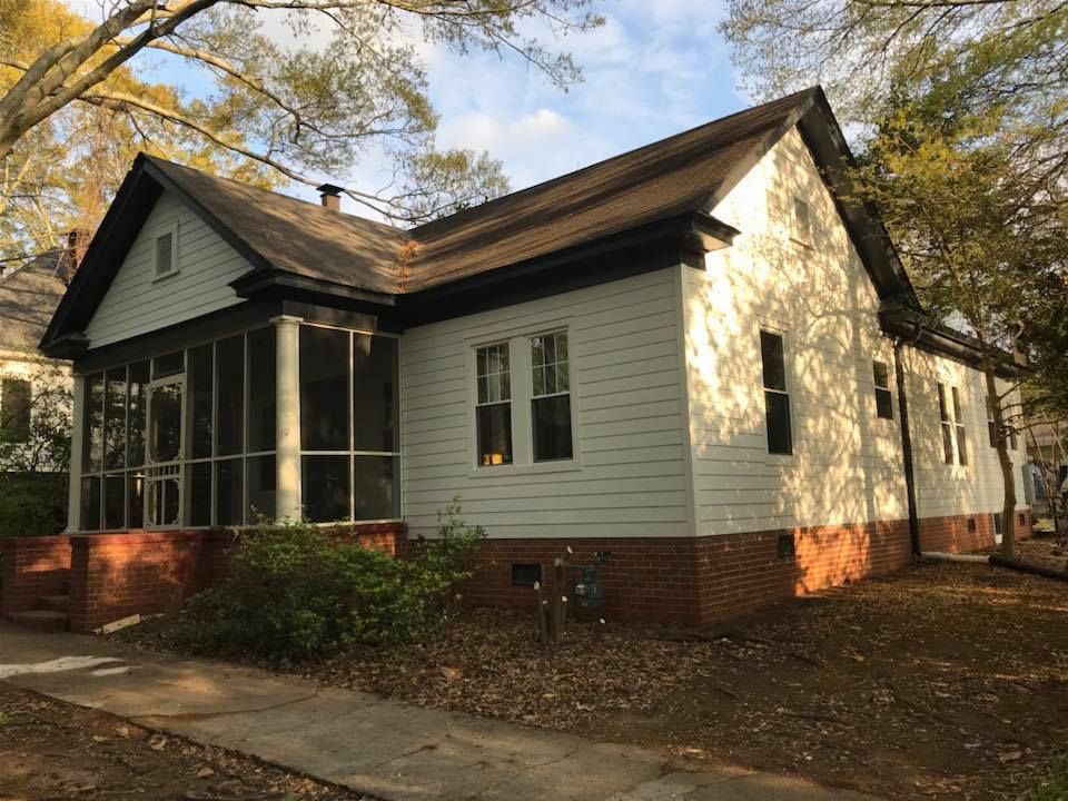 White cottage with a screened-in front porch, brown roof, and brick foundation, surrounded by trees on a sunny day.