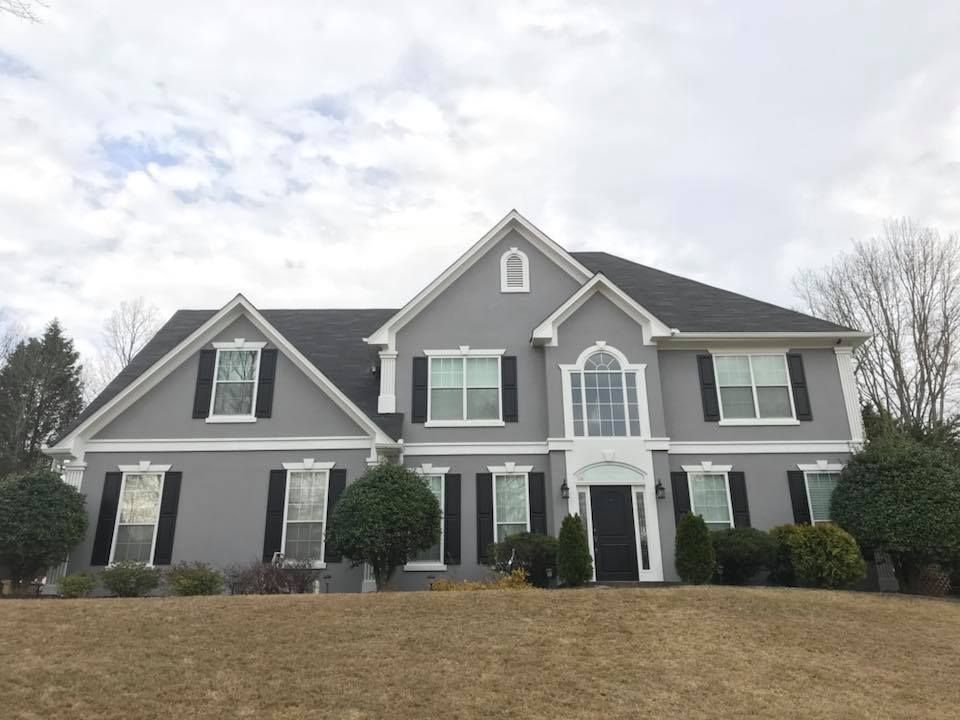 A two-story grey suburban house with white trim, black shutters, and a dark shingled roof under a cloudy sky.