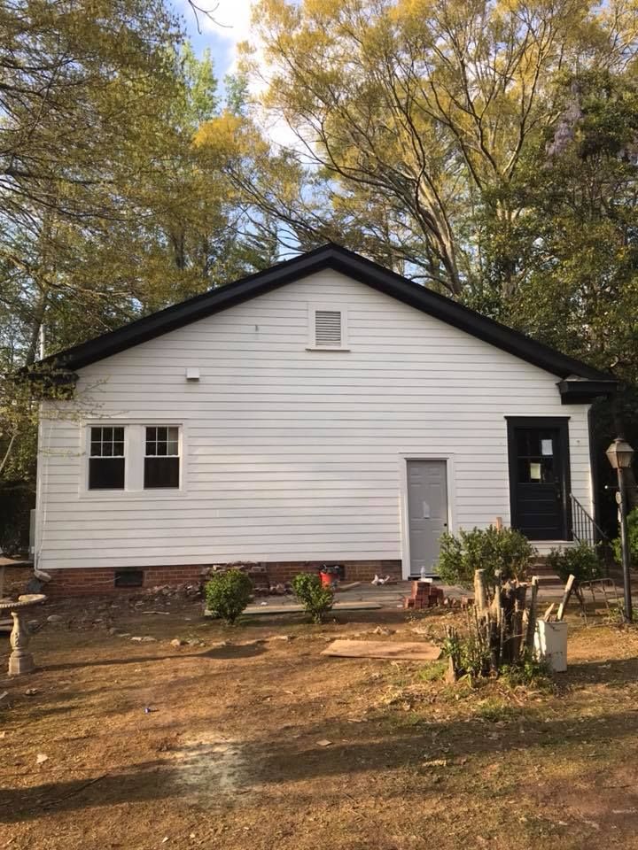 A single-story house with white horizontal siding, black trim, two windows, and two doors, set against a backdrop of trees.
