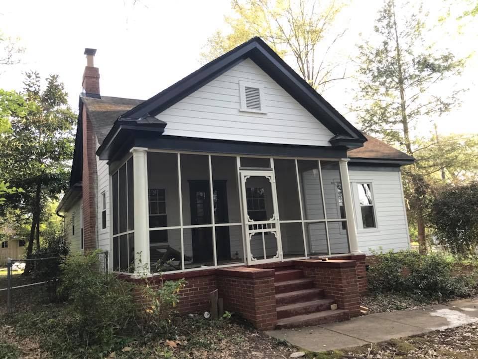 A white cottage-style house with a brick foundation, a screened-in porch, and dark trim, nestled among trees.