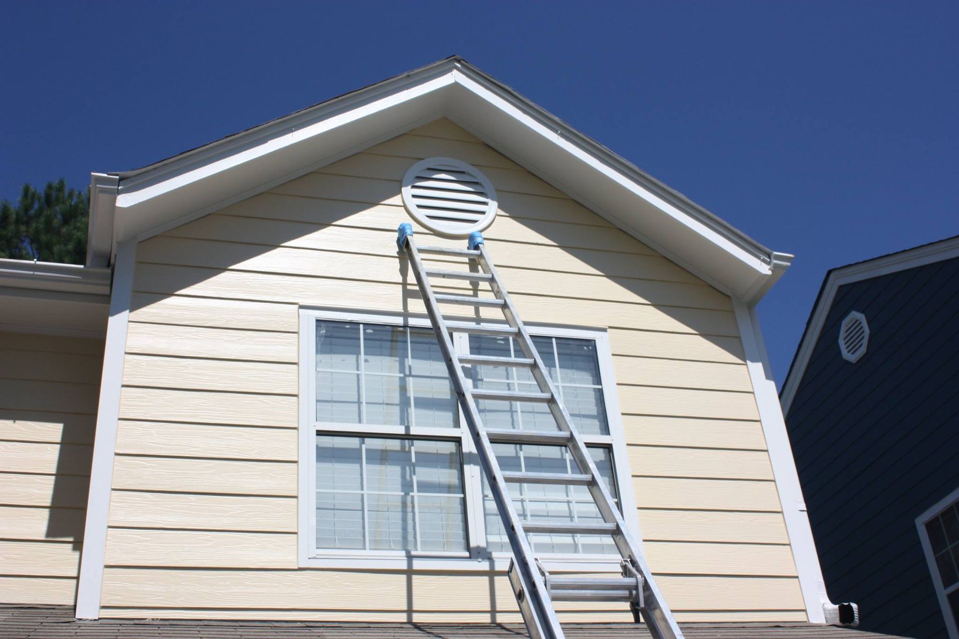 An aluminum extension ladder leans against the exterior wall of a beige house, reaching toward a circular gable vent.