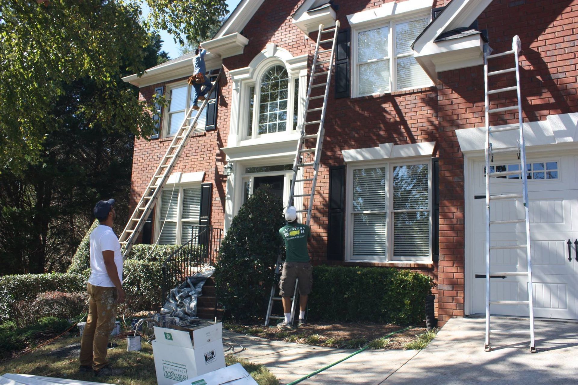 Three people work on the exterior of a red brick house, using ladders to reach the roofline for maintenance.
