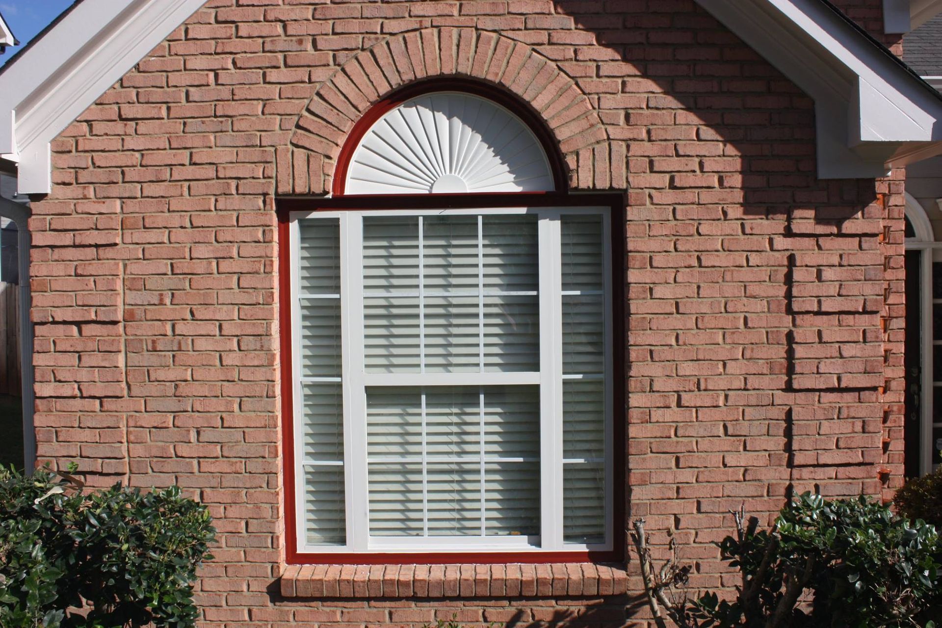 A window with a white frame and blinds, set in a tan brick wall with an arched, sunburst-pattern transom above.