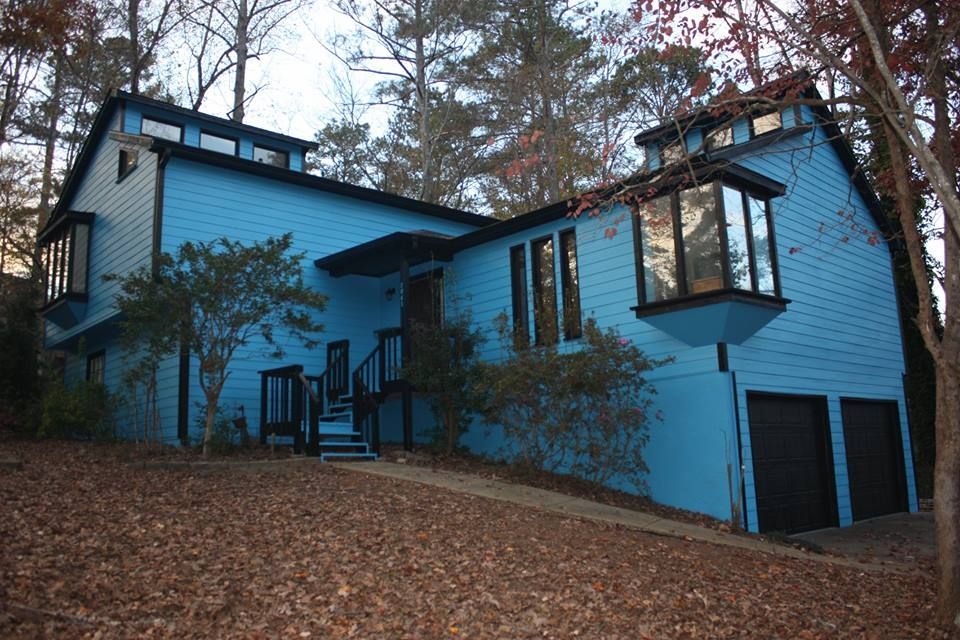 A two-story, bright blue house with dark trim and a two-car garage sits on a sloped, leaf-covered wooded lot.