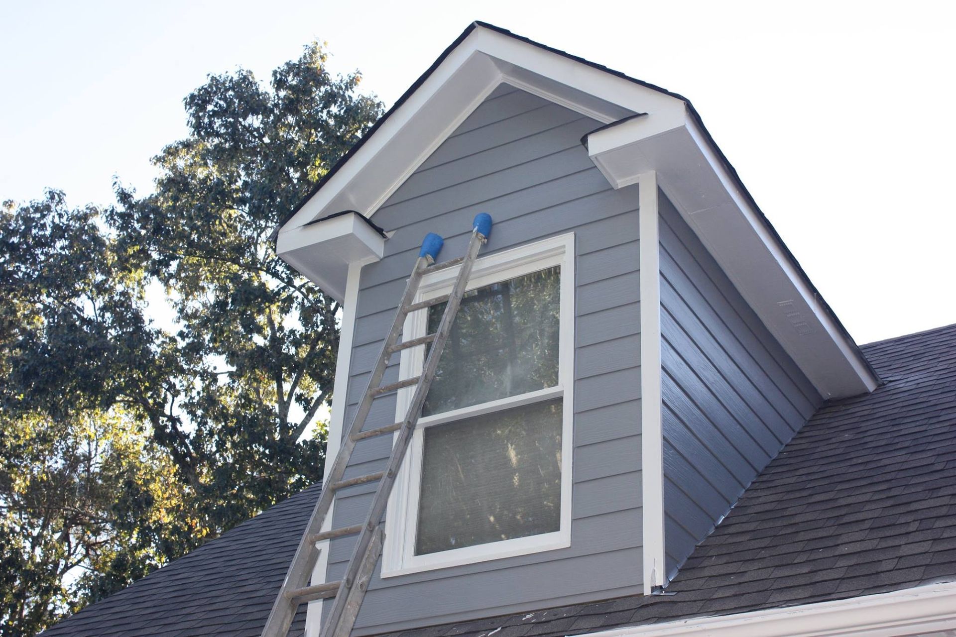 An extension ladder leaning against the side of a gray house, positioned to reach a window on the gabled dormer.