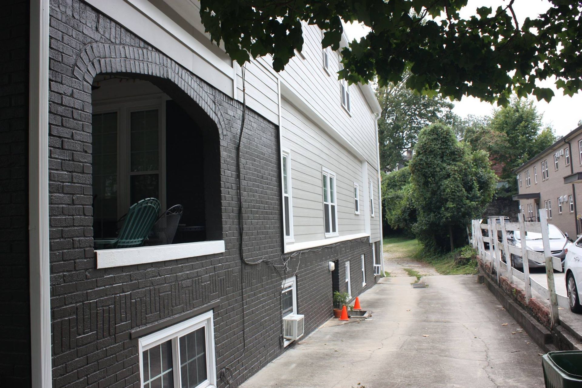 The side view of a house with a dark brick lower level, light siding upper level, and an arched porch along a driveway.