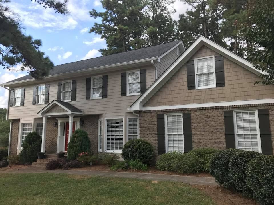 A two-story house with a tan shingle and brick exterior, a front porch, black shutters, and surrounding landscaping.