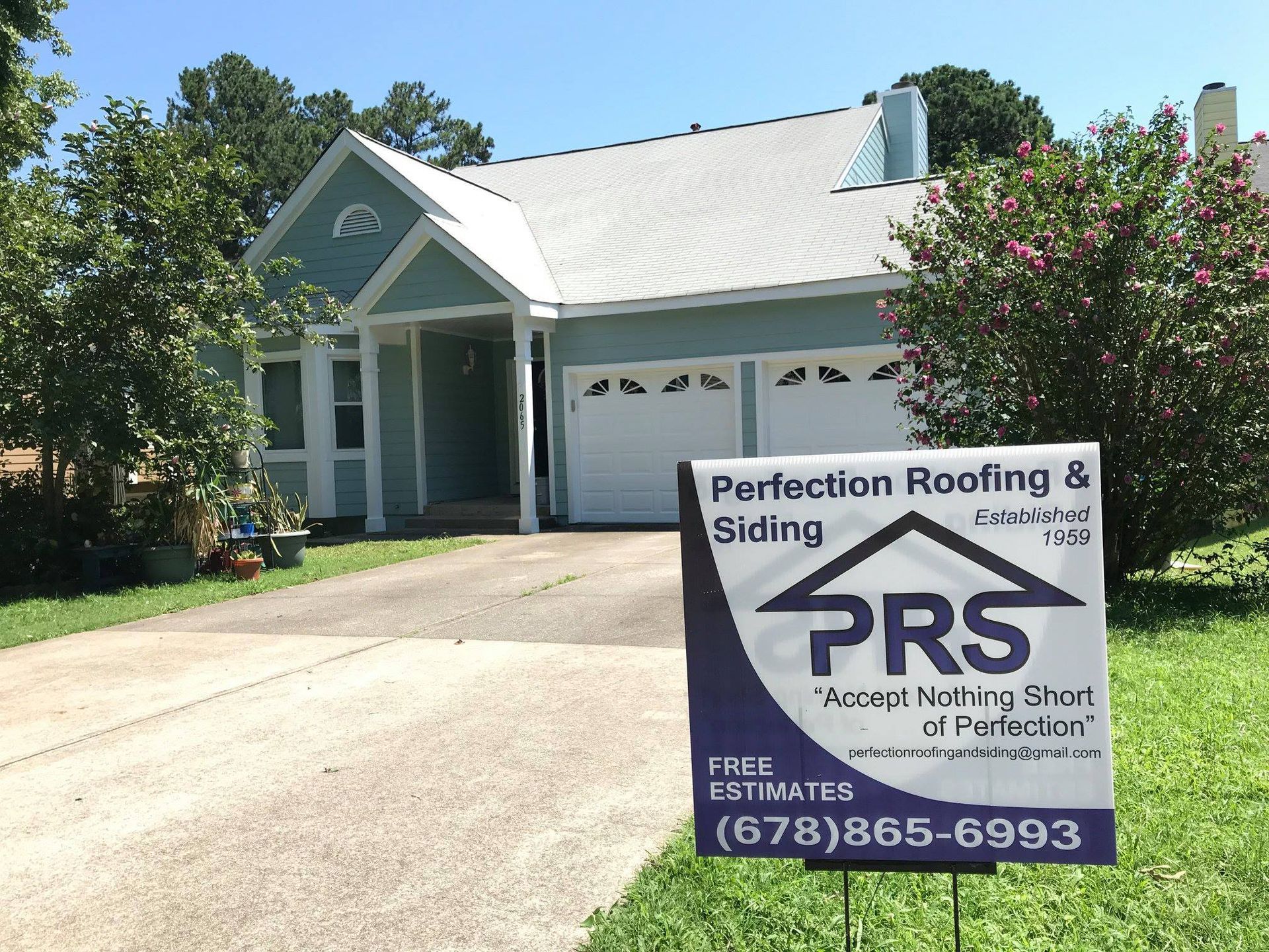 A light blue house with a two-car garage sits behind a promotional lawn sign for Perfection Roofing & Siding.