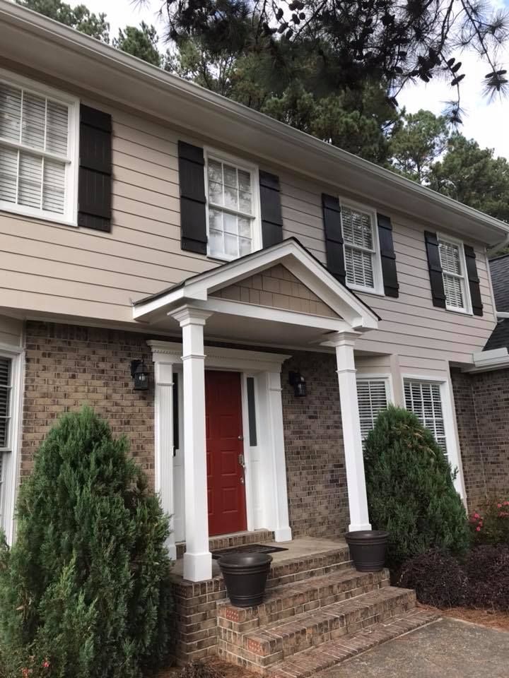 Two-story house with beige siding, brick lower level, black shutters, white columns, and a bright red front door.