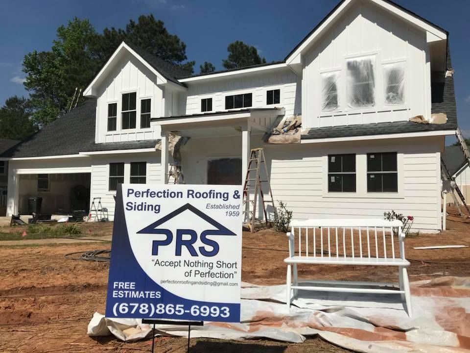 A white, two-story house under construction with a Perfection Roofing & Siding sign and a white bench in the foreground.