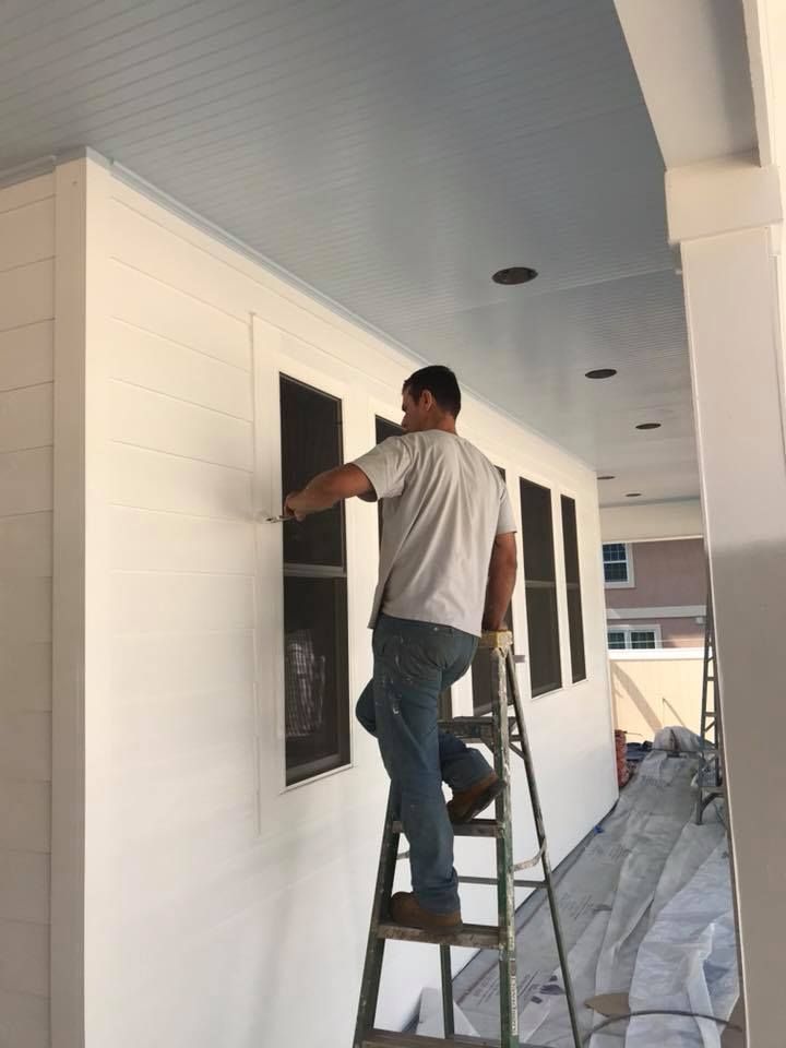 A person on a ladder paints the white trim around a window on the exterior of a home.