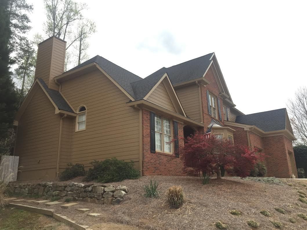 A two-story brick and tan siding house with a dark shingled roof, set on a sloped lot with a stone retaining wall.