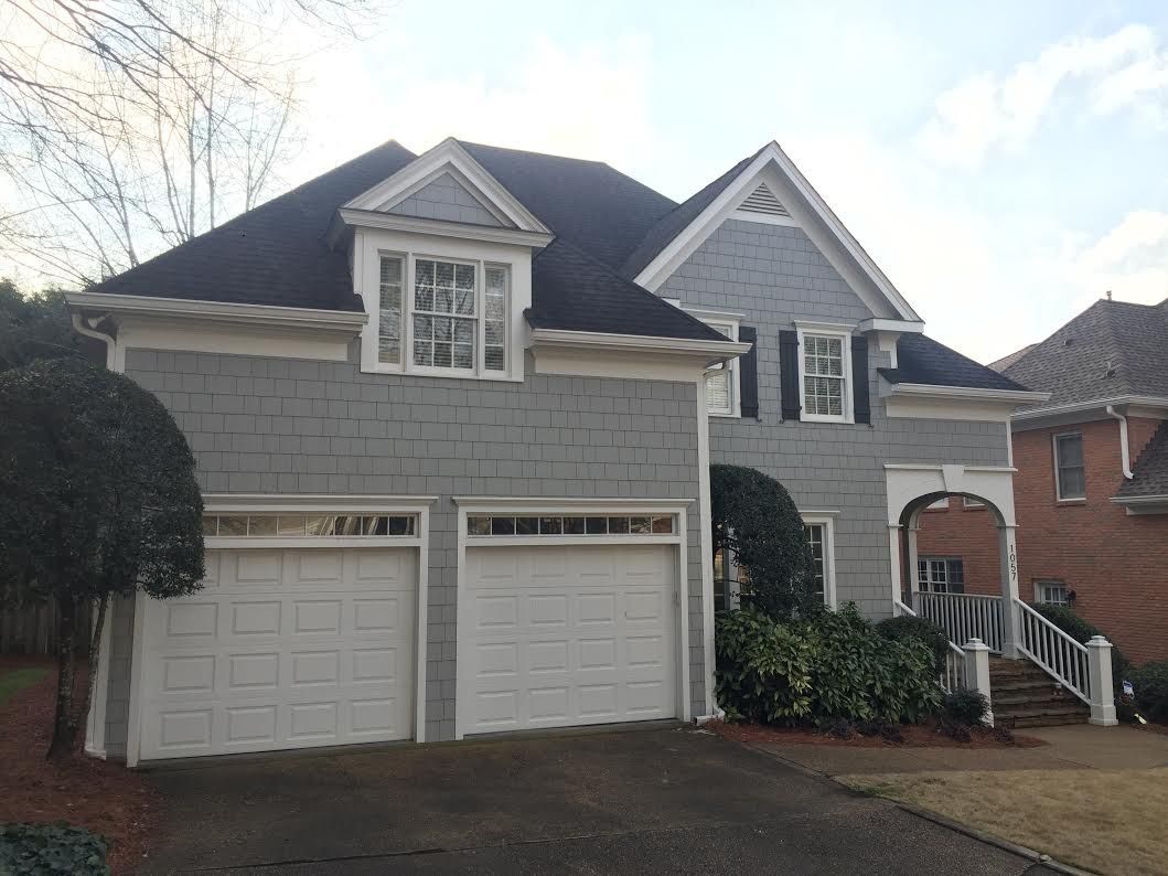 A two-story house with gray shingle siding, white garage doors, a covered porch, and a dark shingled roof.