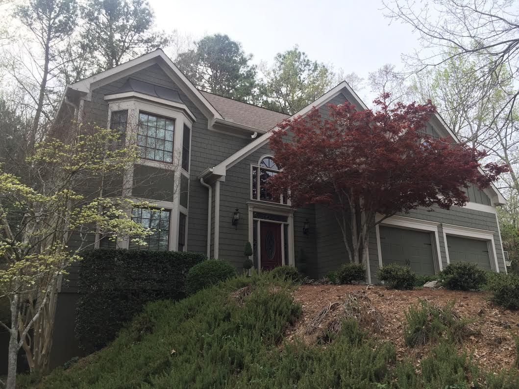 A two-story, grey-sided residential house with a front-facing bay window, a red-leafed tree, and a two-car garage.