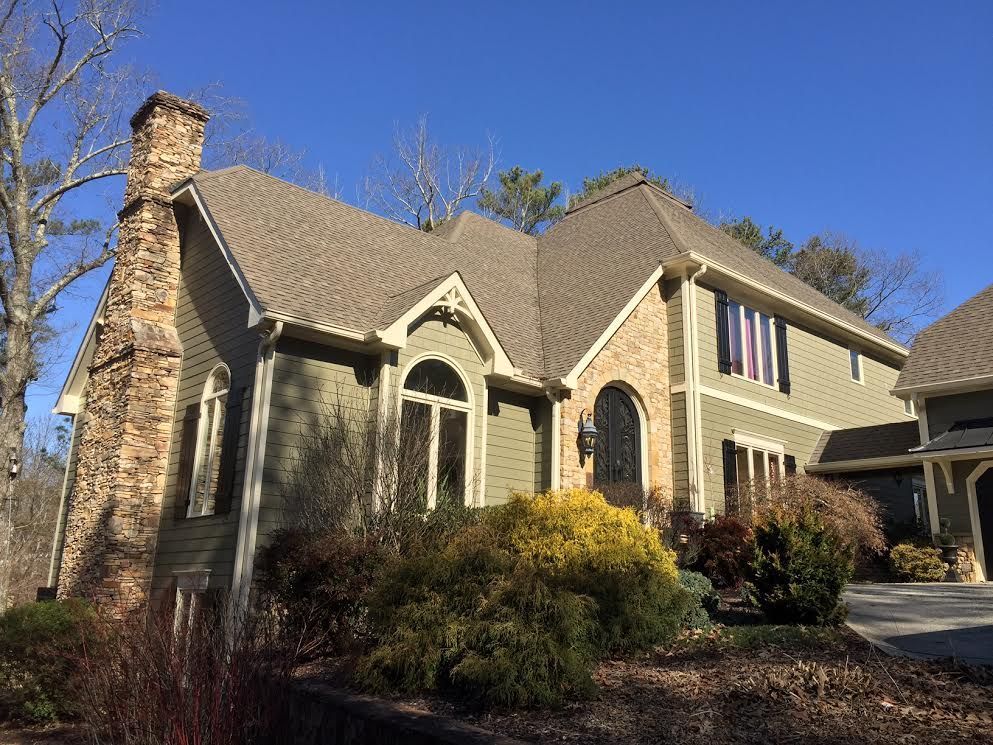 A two-story green house with a stone chimney, arched front entrance, and beige shingled roof under a clear blue sky.