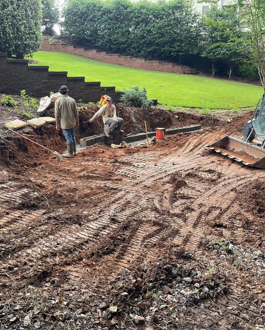 Workers leveling a muddy backyard beside a green lawn, with a trench and construction equipment.