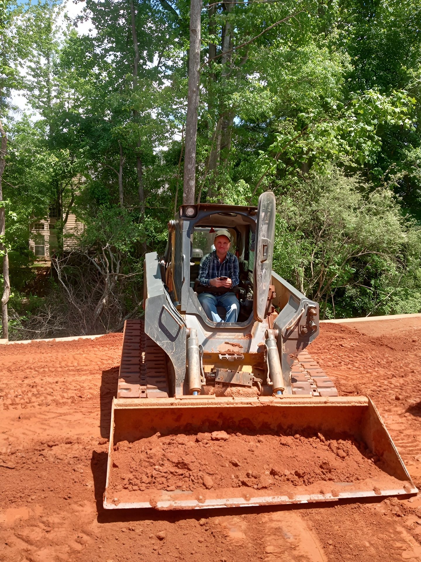 Bulldozer with front blade on a red dirt road, operator seated in the cab, trees in the background