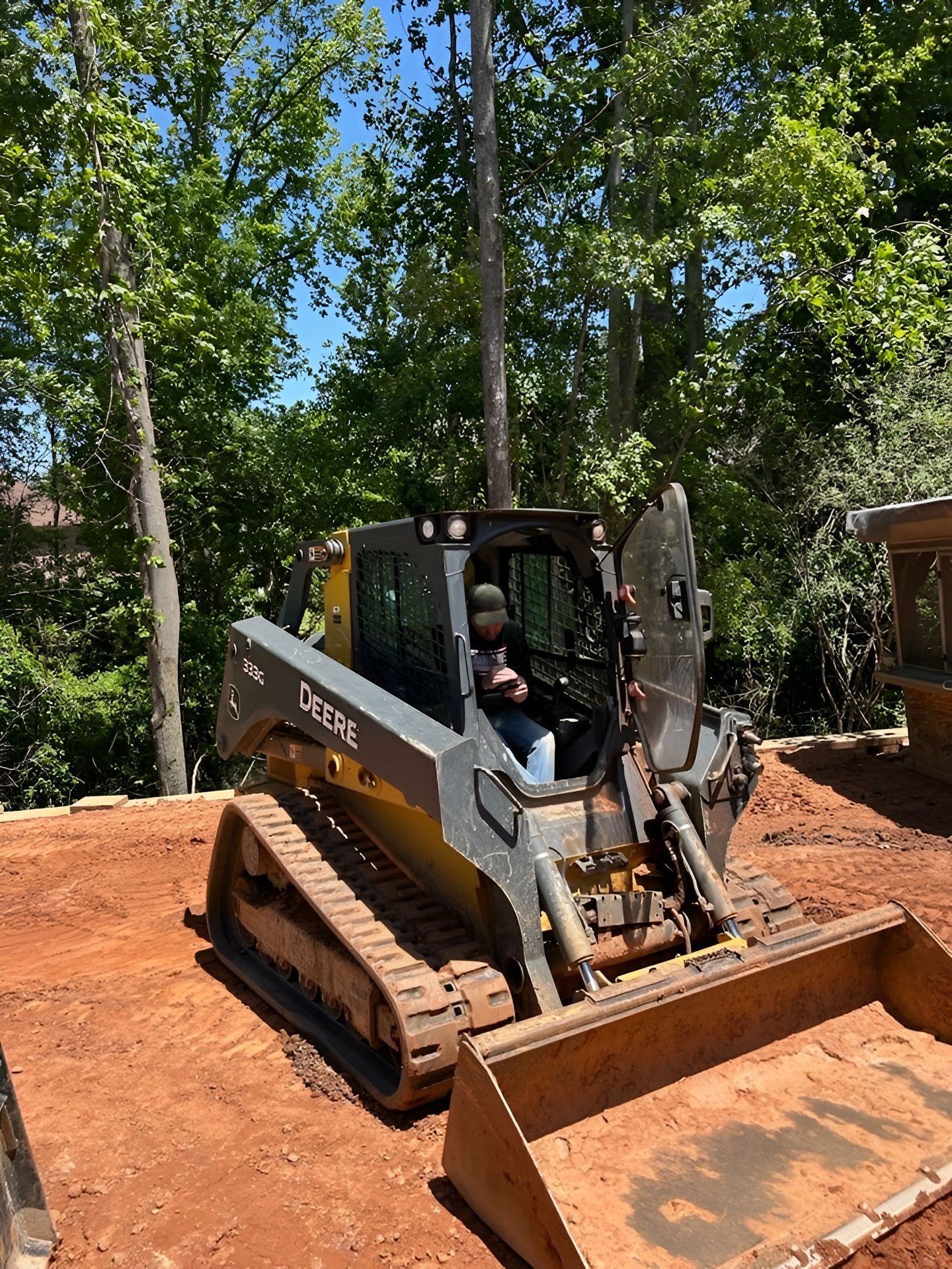 Skid steer with bucket on red dirt at a wooded construction site