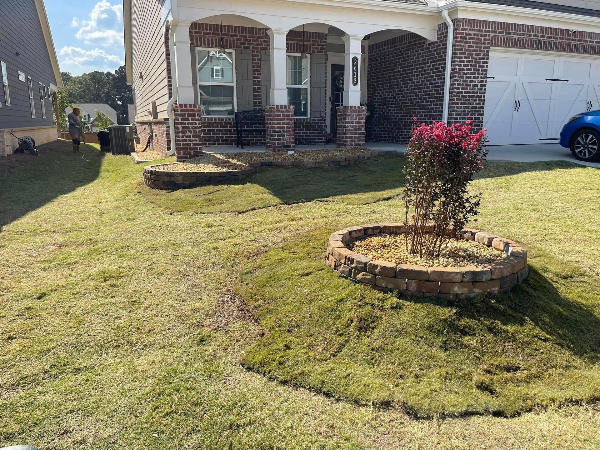 Front yard of a brick house with a porch, green lawn, and a circular flower bed with a red shrub