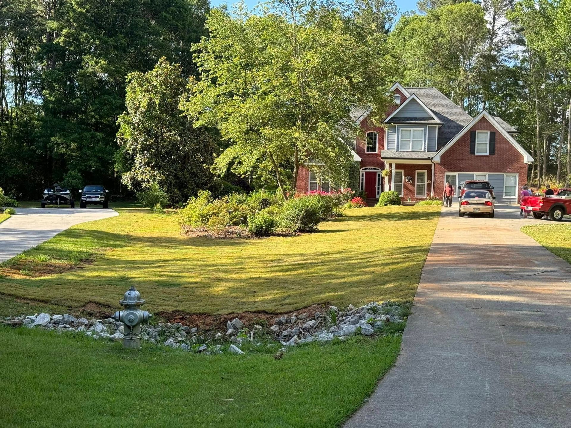 Suburban brick house with a large lawn, driveway, parked cars, and trees on a sunny day