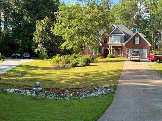 Suburban brick house with a large lawn, driveway, parked cars, and trees on a sunny day