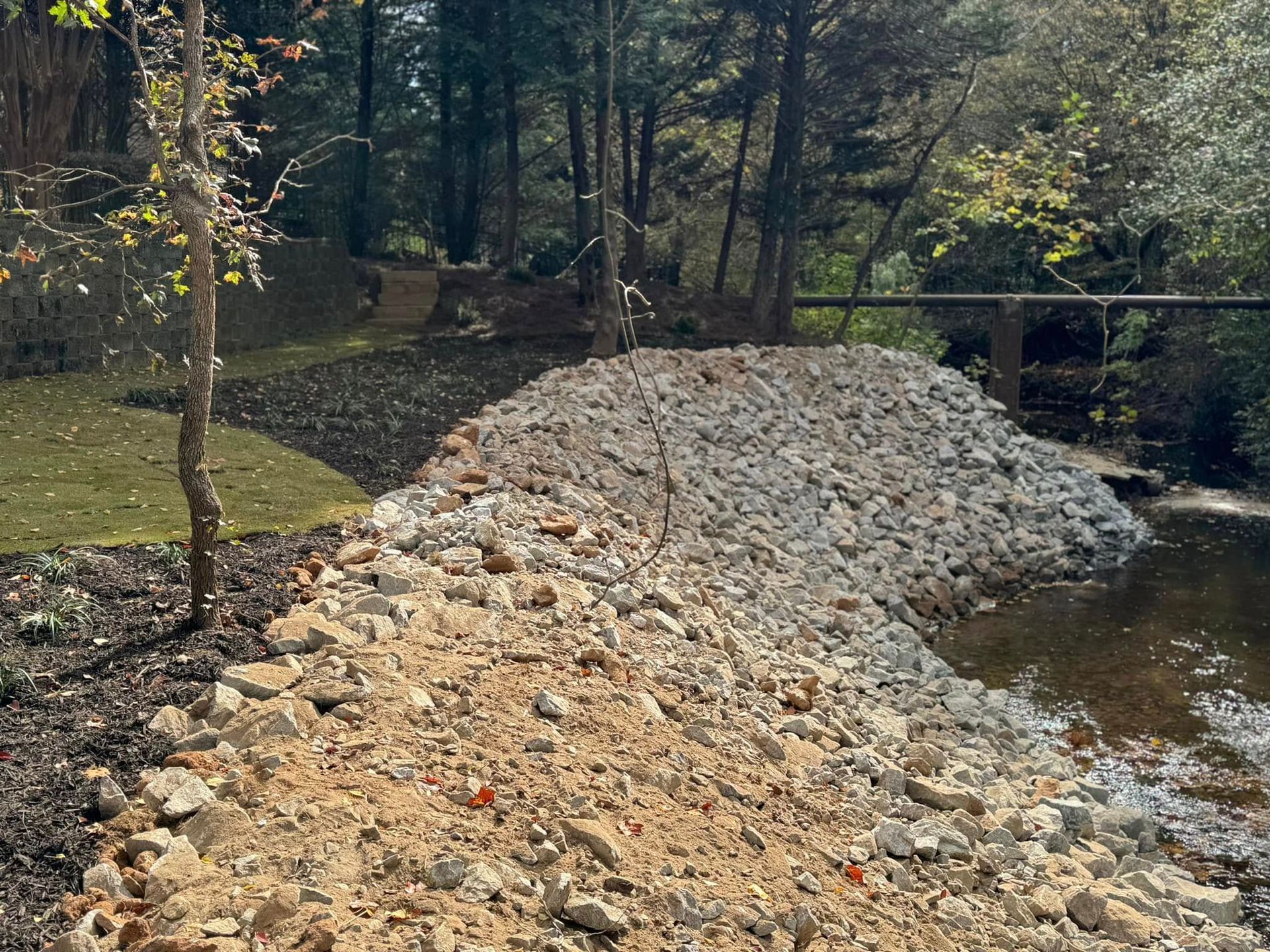 Rock-lined embankment beside a creek in a wooded park, with trees and sunlight in the background