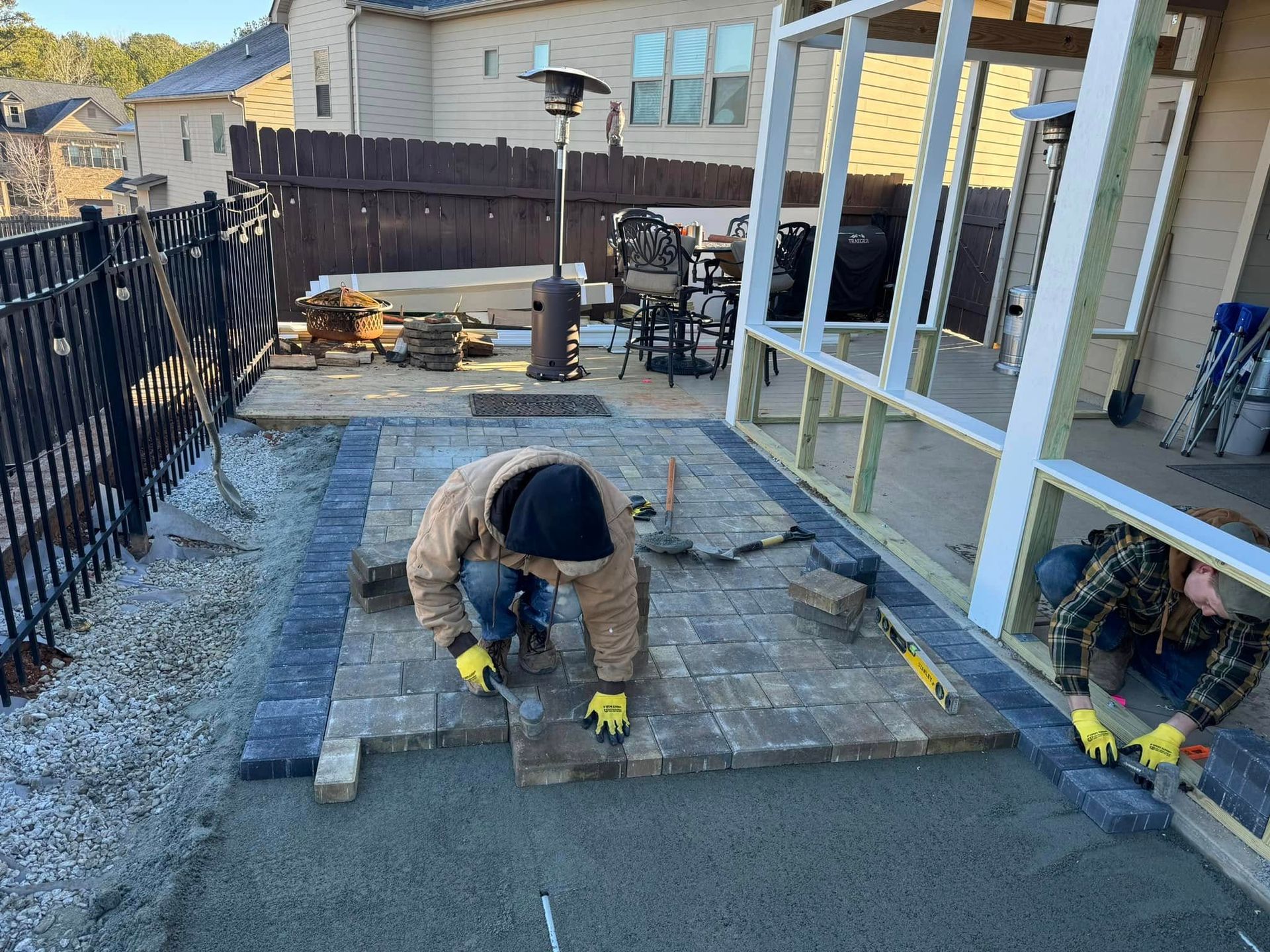 Worker kneeling on a backyard patio, installing stone pavers beside a house and covered porch