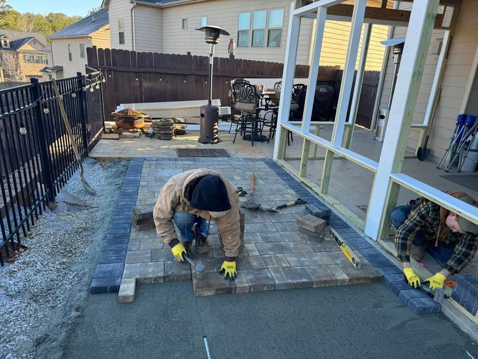 Worker kneeling on a backyard patio, installing stone pavers beside a house and covered porch