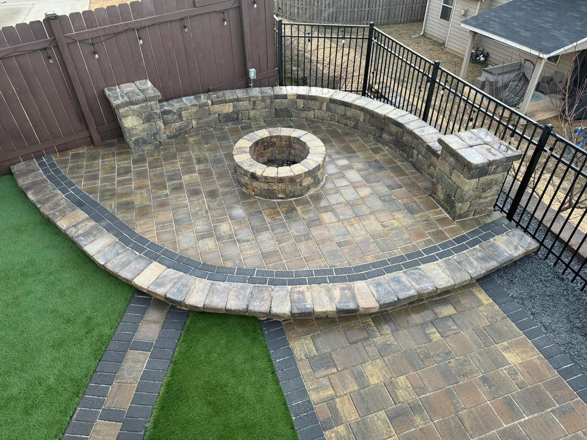 Curved stone patio with a central fire pit, bordered by black fencing and a strip of grass.