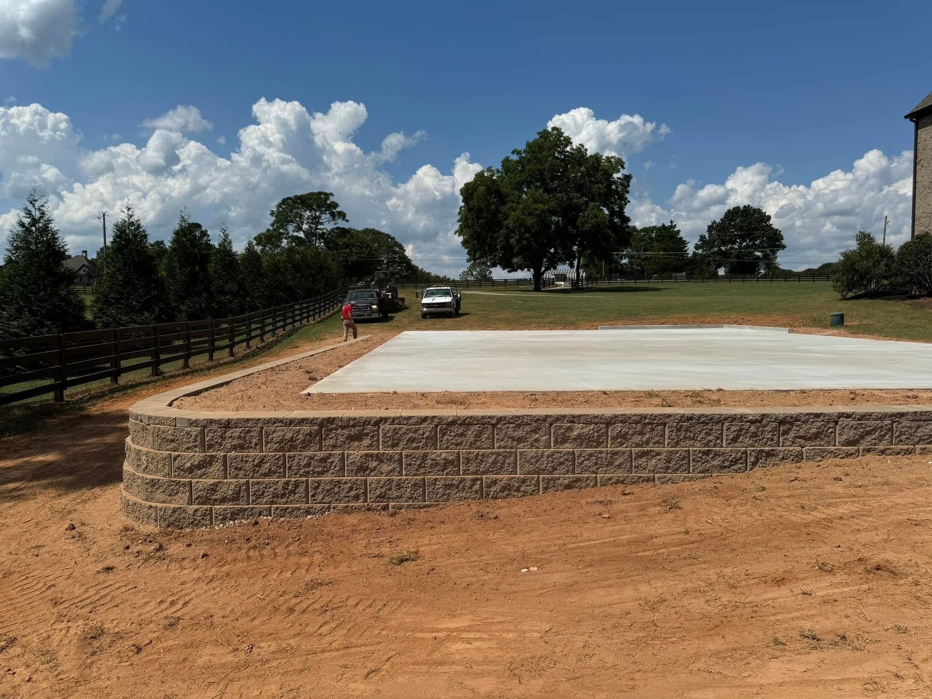 Stone retaining wall around a concrete slab on a dirt lot, with trees and blue sky in the background