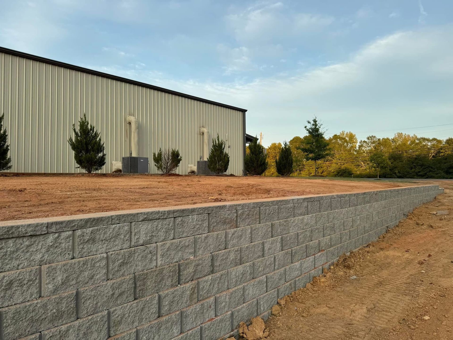 Stone retaining wall beside a tan industrial building with shrubs and trees under a blue sky