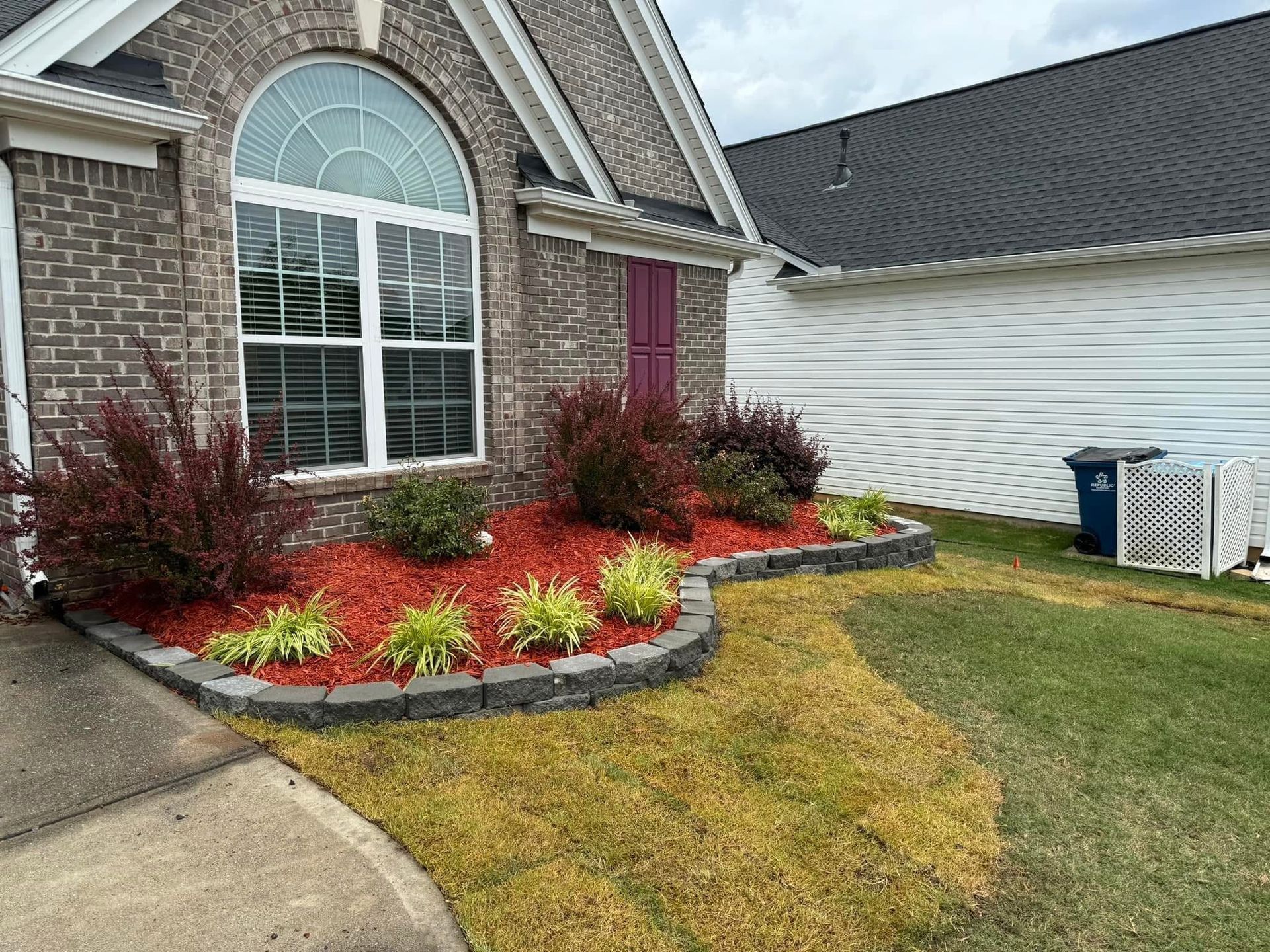 Front yard landscaping with red and yellow shrubs beside a brick house and arched window