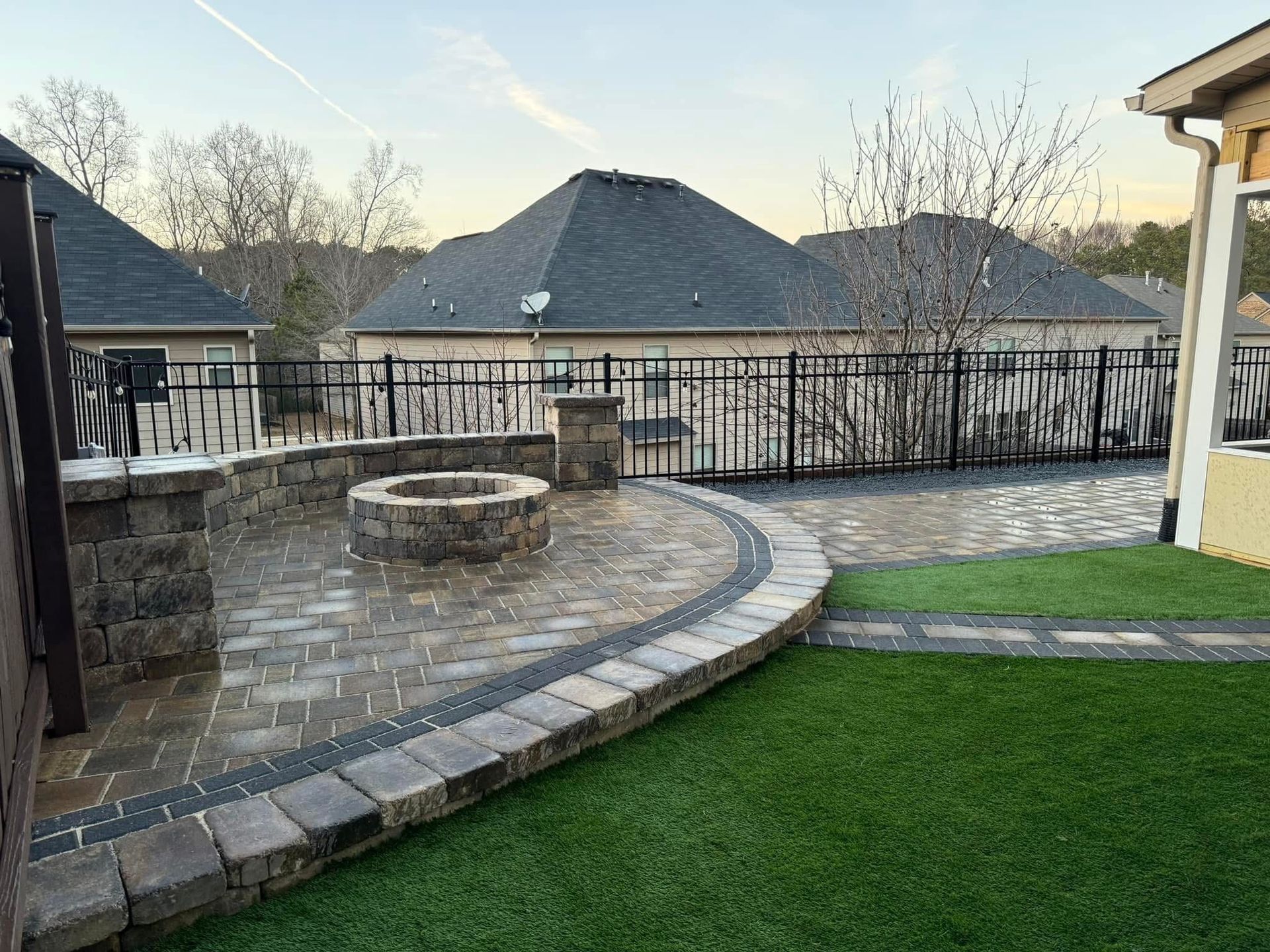 Backyard patio with stone fire pit, curved paving, and green lawn at dusk