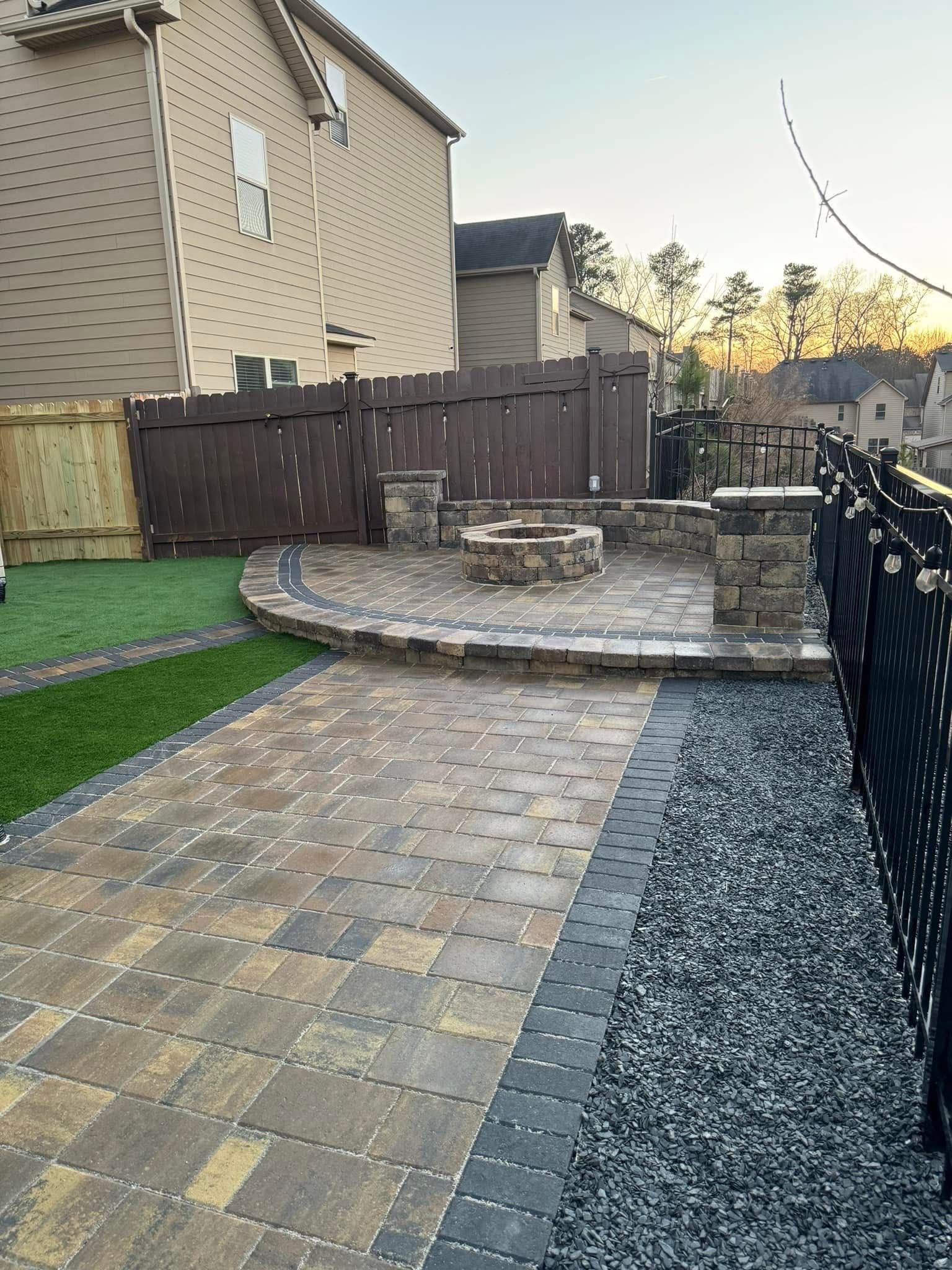 Backyard patio with brick pavers, curved stone fire pit, wooden fence, and gravel border