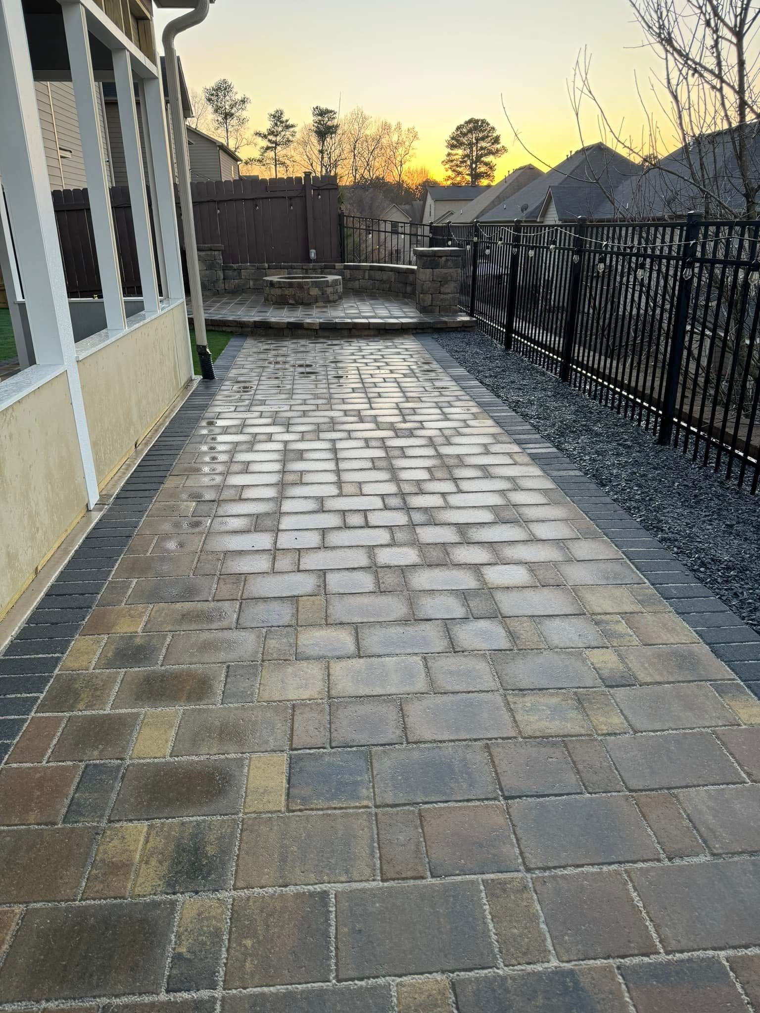 Stone-paver walkway beside a black fence and house at sunset, with wet ground reflecting the sky