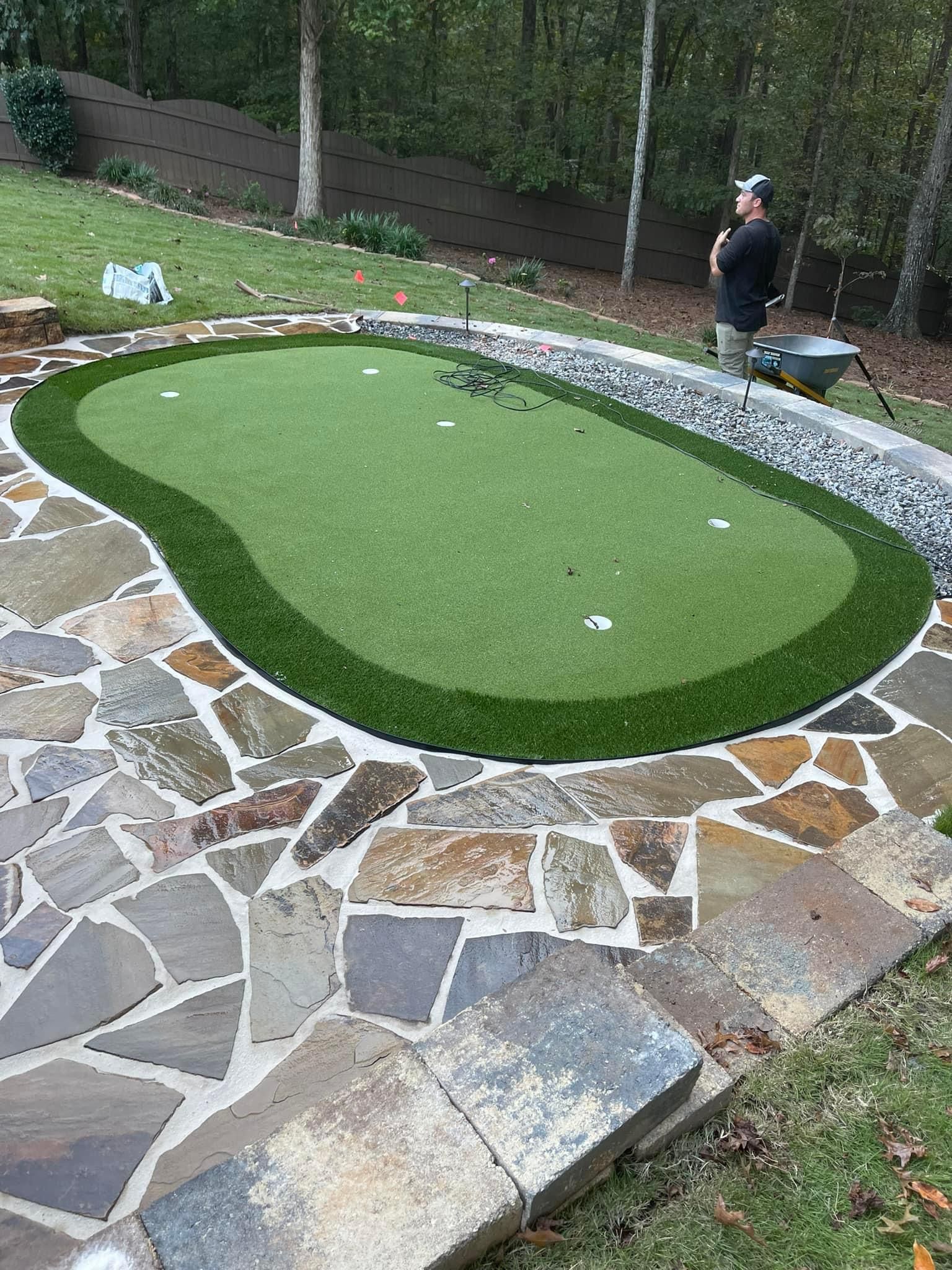 Outdoor putting green with stone border in a backyard; a person stands at the far end.